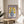 May include: Framed poster print with purple border displaying yellow flowers in a blue polka-dotted vase, positioned on a round wooden side table next to a beige upholstered chair. Natural lighting creates soft shadows on white walls, with green plant leaves visible in the corner of the bright, minimalist room setting.