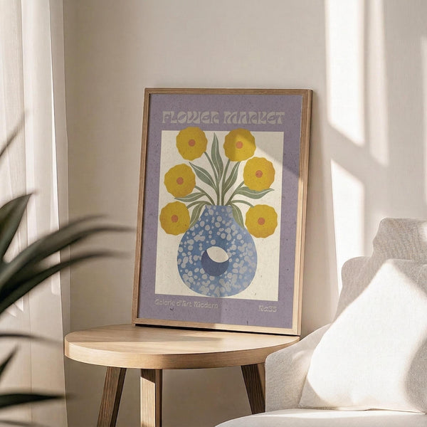 May include: Framed poster print with purple border displaying yellow flowers in a blue polka-dotted vase, positioned on a round wooden side table next to a beige upholstered chair. Natural lighting creates soft shadows on white walls, with green plant leaves visible in the corner of the bright, minimalist room setting.