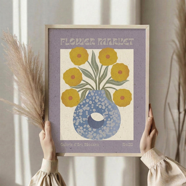 May include: Hands holding a framed vintage-style poster featuring yellow flowers in a blue polka-dotted vase against a cream background with purple border. The poster reads "Flower Market" at top and "Galerie d'Art Moderne No.35" at bottom. Person wears a beige ruffled sleeve top and holds dried pampas grass, photographed against white curtained window background.