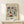 May include: Framed botanical poster with colorful floral pattern in burgundy, teal, and orange tones displayed on light wood shelf against cream wall. Small wrapped gift box with twine sits nearby. Vintage-inspired folk art style features decorative flowers, leaves, and berries in repeating design. Natural wood frame complements warm, minimalist home decor setting.