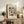 May include: Framed vintage-style botanical poster with colorful folk art flowers in burgundy, teal, and orange tones, displayed on rustic wooden sideboard alongside fern plant, ceramic vase, and books in warm neutral living room setting with cream upholstered chair and brown throw pillow visible.
