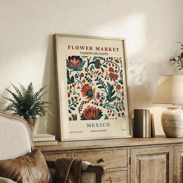 May include: Framed vintage-style botanical poster with colorful folk art flowers in burgundy, teal, and orange tones, displayed on rustic wooden sideboard alongside fern plant, ceramic vase, and books in warm neutral living room setting with cream upholstered chair and brown throw pillow visible.