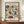 May include: Framed vintage-style botanical poster displayed on wooden credenza against white wainscoted wall. Features colorful folk art floral pattern with orange, burgundy, and teal flowers on cream background. French text at top, "Mexico" at bottom. White ceramic vase and books nearby for scale.