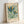 May include: Framed vintage-style botanical poster leaning against white wall. Light wooden frame contains cream-colored print featuring two stylized flowers with blue, teal, orange and green petals surrounded by small decorative blooms. Text reads "FLOWER MARKET" at top and "PARIS" at bottom with museum attribution. Soft natural lighting creates gentle shadows on minimalist background.