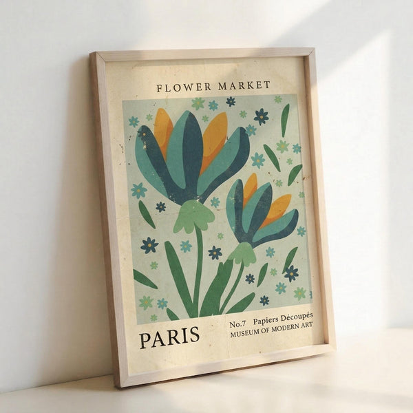 May include: Framed vintage-style botanical poster leaning against white wall. Light wooden frame contains cream-colored print featuring two stylized flowers with blue, teal, orange and green petals surrounded by small decorative blooms. Text reads "FLOWER MARKET" at top and "PARIS" at bottom with museum attribution. Soft natural lighting creates gentle shadows on minimalist background.