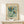 May include: Framed vintage-style botanical poster with stylized blue, teal, and orange flowers on pale green background, displayed on light wood console table next to small wrapped gift with twine and greenery sprig against cream wall