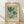 May include: Framed botanical poster print leaning against white wall on light wooden floor. Features stylized flowers with teal and orange petals, green stems, scattered small blooms on pale green background. Text reads "Flower Market Paris" and "Papiers Découpés Museum of Modern Art". Natural wood frame with cream matting, soft lighting creates gentle shadows.