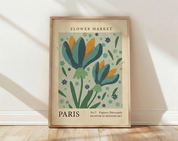 May include: Framed botanical poster print leaning against white wall on light wooden floor. Features stylized flowers with teal and orange petals, green stems, scattered small blooms on pale green background. Text reads "Flower Market Paris" and "Papiers Découpés Museum of Modern Art". Natural wood frame with cream matting, soft lighting creates gentle shadows.