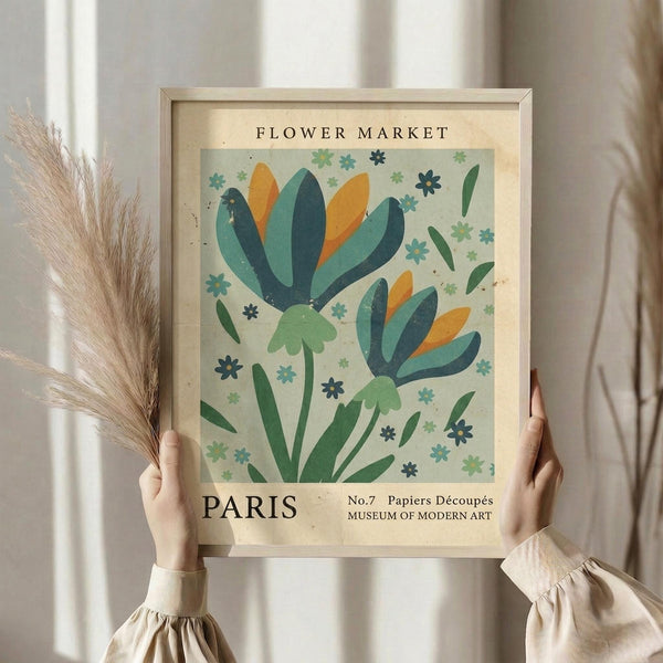 May include: Hands holding a framed vintage-style botanical poster featuring two stylized flowers with teal, orange, and green petals surrounded by small decorative blooms. The aged cream-colored print reads "Flower Market Paris" and "Museum of Modern Art" with dried pampas grass visible beside it against soft natural lighting.