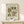 May include: Vintage botanical art print in wooden frame displaying ornate floral patterns in olive green, rust orange, and golden yellow colors. Features decorative flowers, leaves, and scrollwork in folk art style with "Flower Market" and "Santorini" text. Frame sits on light wood surface against cream wall with small wrapped gift package beside it.