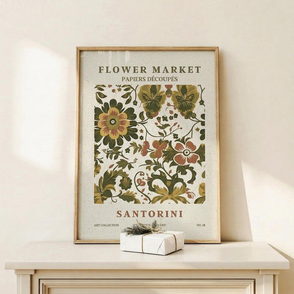 May include: Vintage botanical art print in wooden frame displaying ornate floral patterns in olive green, rust orange, and golden yellow colors. Features decorative flowers, leaves, and scrollwork in folk art style with "Flower Market" and "Santorini" text. Frame sits on light wood surface against cream wall with small wrapped gift package beside it.