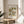 May include: Framed vintage botanical poster with ornate floral pattern in green, brown and cream tones displayed on round wooden side table next to beige upholstered chair. Natural sunlight streams across neutral wall creating soft shadows. Live plant visible in corner of minimalist room setting.