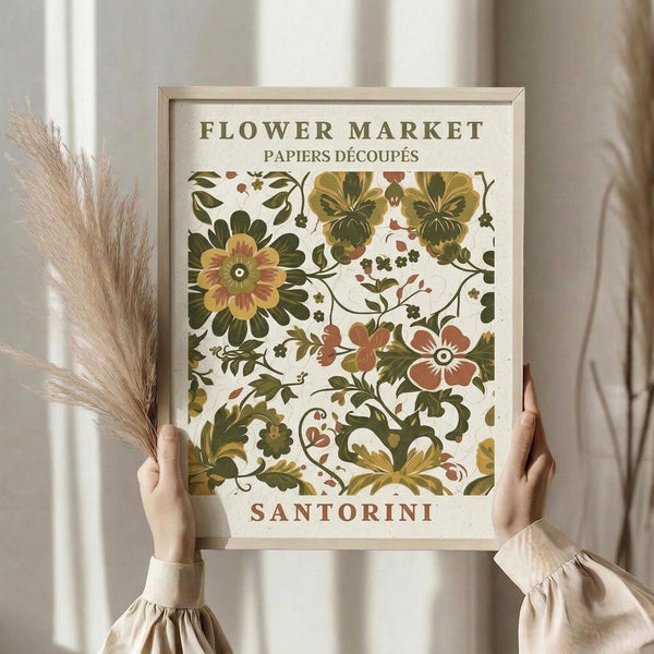May include: A person in cream clothing holding a framed botanical poster featuring vintage-style flowers in green, yellow, and coral tones with "FLOWER MARKET" and "SANTORINI" text. The scene shows dried pampas grass beside them against soft white curtains in natural lighting.