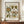 May include: Framed vintage-style botanical poster with ornate floral pattern in greens, yellows, and rust colors displayed on wooden sideboard next to white ceramic vase and stacked books. Features decorative flowers and leaves in folk art style with text headers and exhibition details on cream background in dark wood frame.