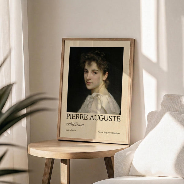 May include: Framed exhibition poster featuring a classical portrait of a young woman with dark curly hair in a white dress against black background. Wooden frame with cream matting displays on round wooden side table next to beige upholstered chair. Soft natural lighting creates shadows on neutral wall. Green plant leaves visible in corner.
