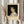 May include: Hands holding framed vintage-style exhibition poster featuring classical portrait painting of young woman with curly dark hair in white dress against black background. Cream matting surrounds artwork with "PIERRE AUGUSTE exhibition" text below. Dried pampas grass visible to left, soft neutral curtains in background create elegant room setting.