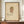 May include: Framed vintage botanical illustration of a fan-shaped coral or sea fan specimen in muted browns and greens on aged cream paper, displayed on a wooden sideboard beside white ceramic vase and stacked books in a room with cream wainscoting and white walls.