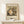 May include: Framed art print of Gustav Klimt's Baby (Cradle) painting featuring a swirling composition of colorful fabrics and patterns surrounding a pale infant figure, displayed in thin wooden frame on light wood surface beside small white wrapped gift with greenery, against cream wall with soft natural lighting