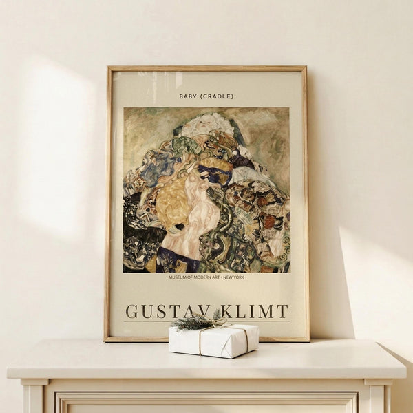 May include: Framed art print of Gustav Klimt's Baby (Cradle) painting featuring a swirling composition of colorful fabrics and patterns surrounding a pale infant figure, displayed in thin wooden frame on light wood surface beside small white wrapped gift with greenery, against cream wall with soft natural lighting