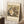 May include: Framed reproduction poster of Gustav Klimt's 1917 painting "Baby (Cradle)" leaning against white wall in cozy room setting. Artwork shows swirling, colorful composition with mother and infant in Klimt's signature decorative style with ornate patterns in greens, golds, and earth tones. Cream matted print in light wood frame positioned on white textured rug beside wooden stool with coffee cup and beige throw blanket.