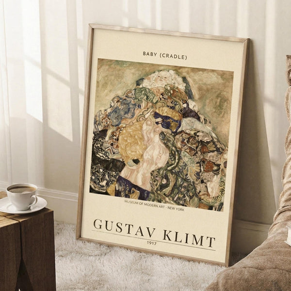 May include: Framed reproduction poster of Gustav Klimt's 1917 painting "Baby (Cradle)" leaning against white wall in cozy room setting. Artwork shows swirling, colorful composition with mother and infant in Klimt's signature decorative style with ornate patterns in greens, golds, and earth tones. Cream matted print in light wood frame positioned on white textured rug beside wooden stool with coffee cup and beige throw blanket.