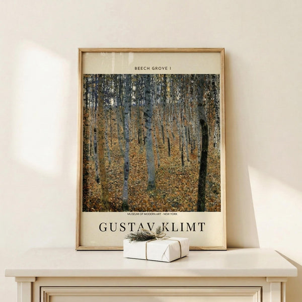 May include: Framed museum poster reproduction of Gustav Klimt's Beech Grove I painting showing tall birch trees with white bark in an autumn forest with golden fallen leaves covering the ground. The poster features cream colored matting with black text identifying the artwork title and Museum of Modern Art New York. Displayed in light wood frame on white wooden surface alongside small white wrapped gift box with natural twine and dried sprig.
