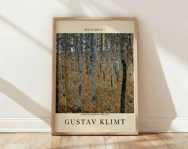 May include: Framed museum poster print of Gustav Klimt's Beech Grove I artwork leaning against white wall on light wood floor. The reproduction shows a dense forest of tall birch trees with white bark and golden autumn foliage covering the ground. The print features cream matting with black text identifying the artwork title, artist name, and Museum of Modern Art New York. Natural lighting creates soft shadows across the minimal room setting with white baseboards visible.
