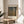 May include: Framed art print of Gustav Klimt's Beech Grove I painting featuring golden autumn birch trees in a forest, displayed on a round wooden side table next to a beige upholstered chair. Sunlight streams across white walls creating soft shadows, with a green plant visible in the corner.