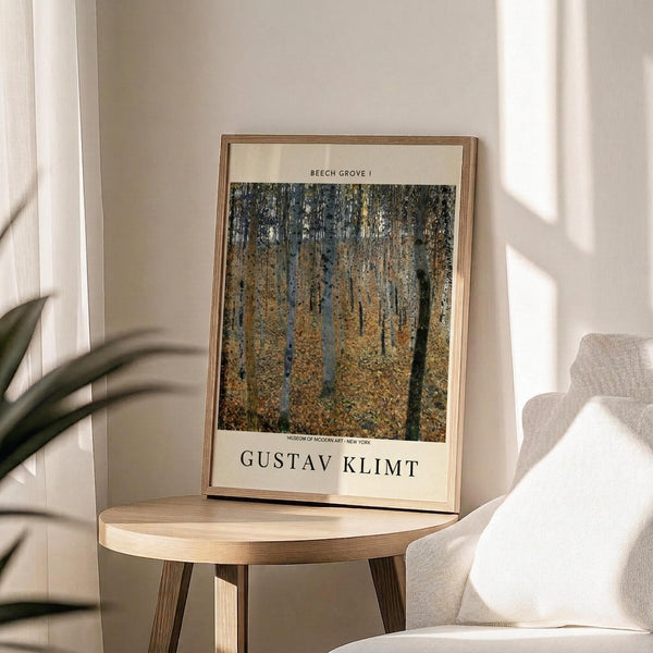 May include: Framed art print of Gustav Klimt's Beech Grove I painting featuring golden autumn birch trees in a forest, displayed on a round wooden side table next to a beige upholstered chair. Sunlight streams across white walls creating soft shadows, with a green plant visible in the corner.