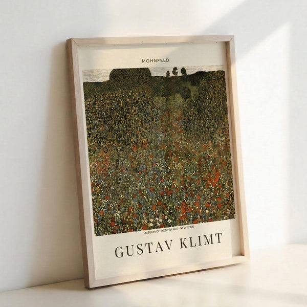 May include: Framed art print of Gustav Klimt's "Mohnfeld" showing a vibrant wildflower meadow with red poppies and colorful blooms covering rolling hills. The poster features rich greens, reds, and yellows in Klimt's distinctive pointillist style, with "GUSTAV KLIMT" and "MUSEUM OF MODERN ART - NEW YORK" text below the image. Natural wood frame leans against white wall on light wooden surface.