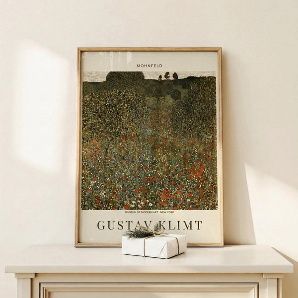 May include: Framed poster print of Gustav Klimt's "Mohnfeld" showing a vibrant wildflower meadow in pointillist style with red poppies and mixed blooms against rolling green hills. The artwork features dense, textured brushwork in earthy greens, reds, and yellows. Natural wood frame sits on light wooden shelf beside small white wrapped gift with greenery sprig against cream wall.