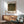 May include: Framed Gustav Klimt poster print showing a colorful wildflower field with green hills and trees in the background, displayed above a wooden mid-century modern credenza with a white ceramic table lamp. A sage green upholstered armchair with wooden arms sits nearby on a wooden floor, with natural sunlight casting geometric shadows across the white wall.