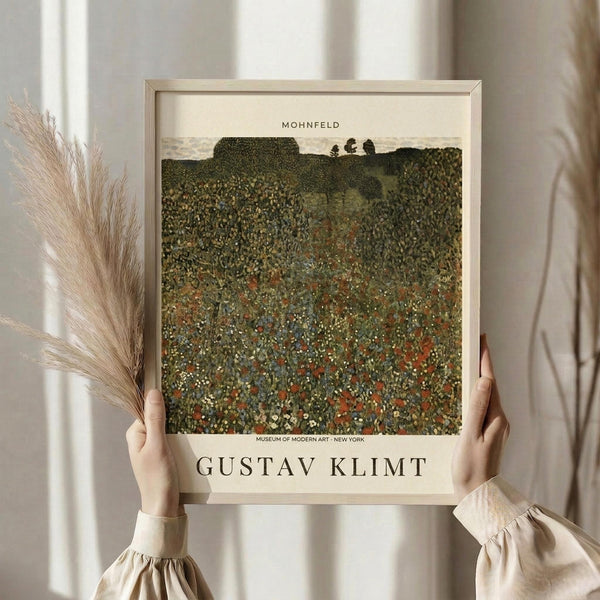 May include: Person in beige clothing holding a framed art poster featuring Gustav Klimt's pointillist landscape painting of a wildflower meadow in greens, reds, and earth tones. The cream-colored frame displays "MOHNFELD" at top and "GUSTAV KLIMT" at bottom with museum attribution. Dried pampas grass visible on left side against soft white curtained background.
