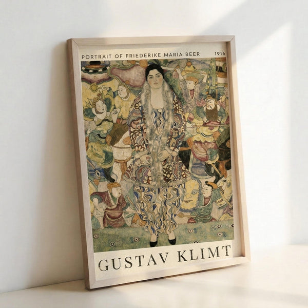 May include: Framed art print leaning against white wall showing Gustav Klimt's Portrait of Friederike Maria Beer from 1916. The colorful painting features a woman with long dark hair in an ornate patterned dress surrounded by stylized figures and decorative motifs in Klimt's distinctive Art Nouveau style. Print has white border with artist name at bottom, displayed in light wood frame on white surface.