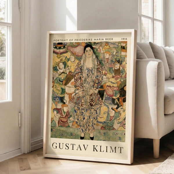 May include: Framed poster leaning against white wall next to beige sofa in bright living room. Artwork shows colorful portrait of woman with long dark hair surrounded by ornate floral and decorative patterns in greens, oranges, and earth tones. Text reads "Portrait of Friederike Maria Beer 1916" and "Gustav Klimt" in black letters on cream background below image.