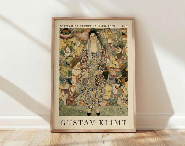 May include: Framed art print displaying Gustav Klimt's 1916 Portrait of Friederike Maria Beer, featuring a woman with long dark hair in an ornate patterned dress surrounded by colorful decorative figures and motifs in Klimt's distinctive Art Nouveau style. The poster has cream matting with black text identifying the artwork and artist, housed in a light wood frame, leaning against white walls on light wooden flooring with soft natural lighting.