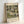 May include: Framed art print in light wooden frame leaning against white wall. Shows Gustav Klimt's painting of an Italian lakeside village with white buildings, tall dark cypress trees, domed church, and water in foreground. Artwork features muted greens, golds, and cream tones in Klimt's decorative style. Text reads "Kirche in Cassone" at top and "Gustav Klimt" with "Museum of Modern Art New York" at bottom on cream background.