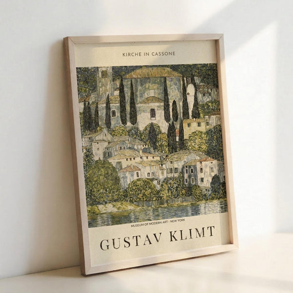 May include: Framed art print in light wooden frame leaning against white wall. Shows Gustav Klimt's painting of an Italian lakeside village with white buildings, tall dark cypress trees, domed church, and water in foreground. Artwork features muted greens, golds, and cream tones in Klimt's decorative style. Text reads "Kirche in Cassone" at top and "Gustav Klimt" with "Museum of Modern Art New York" at bottom on cream background.