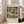 May include: A framed reproduction of Gustav Klimt's "Kirche in Cassone" artwork displayed in a bright living room setting. The poster shows a lakeside village scene with Mediterranean-style buildings, tall cypress trees, and a prominent church with golden domes, rendered in Klimt's characteristic muted greens, golds, and earth tones. The print has cream-colored text identifying the artwork and Museum of Modern Art, housed in a light wooden frame leaning against white walls near a beige so