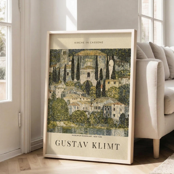 May include: A framed reproduction of Gustav Klimt's "Kirche in Cassone" artwork displayed in a bright living room setting. The poster shows a lakeside village scene with Mediterranean-style buildings, tall cypress trees, and a prominent church with golden domes, rendered in Klimt's characteristic muted greens, golds, and earth tones. The print has cream-colored text identifying the artwork and Museum of Modern Art, housed in a light wooden frame leaning against white walls near a beige so