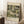 May include: A framed art print leaning against a white wall in a cozy room setting. The poster features a scenic landscape painting with Mediterranean-style buildings, cypress trees, and a church dome in earthy greens, yellows and whites. The artwork has cream-colored borders with text at top and bottom. A coffee cup sits on a wooden surface nearby, with a textured rug and neutral bedding visible in the soft, natural lighting.