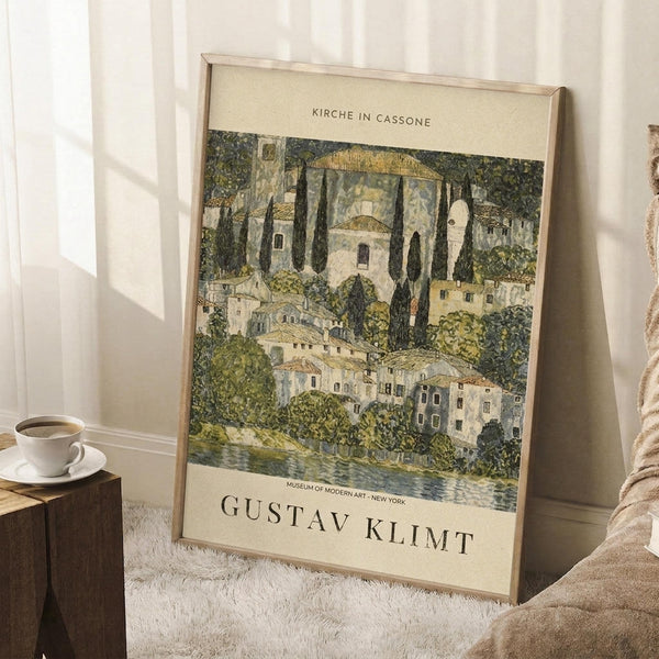May include: A framed art print leaning against a white wall in a cozy room setting. The poster features a scenic landscape painting with Mediterranean-style buildings, cypress trees, and a church dome in earthy greens, yellows and whites. The artwork has cream-colored borders with text at top and bottom. A coffee cup sits on a wooden surface nearby, with a textured rug and neutral bedding visible in the soft, natural lighting.