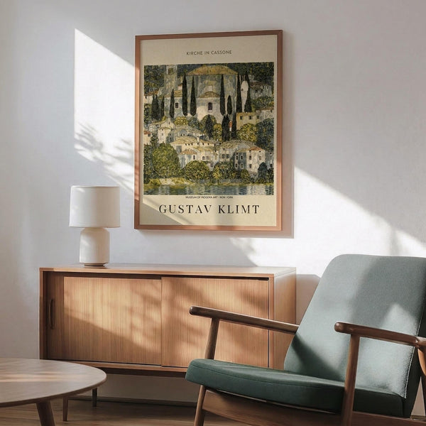May include: Gustav Klimt art print in wooden frame hung above mid-century modern credenza with white table lamp. Sage green upholstered armchair with wood arms sits nearby. Print shows Mediterranean church and village buildings surrounded by cypress trees and foliage in Klimt's distinctive style with muted greens, yellows and earth tones. Bright natural light creates dramatic shadows across white wall in modern living room setting.