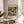 May include: Framed art print featuring a colorful landscape painting with Mediterranean buildings, cypress trees, and waterfront architecture in green, gold, and earth tones. The poster displays museum exhibition text and artist name at bottom. Shown in thin wood frame on round wooden side table next to beige upholstered chair with decorative pillow, green plant leaves visible, soft natural lighting creating shadows on neutral wall.