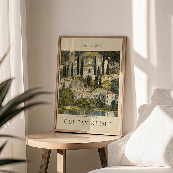 May include: Framed art print featuring a colorful landscape painting with Mediterranean buildings, cypress trees, and waterfront architecture in green, gold, and earth tones. The poster displays museum exhibition text and artist name at bottom. Shown in thin wood frame on round wooden side table next to beige upholstered chair with decorative pillow, green plant leaves visible, soft natural lighting creating shadows on neutral wall.