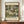 May include: Framed reproduction of Gustav Klimt's "Kirche in Cassone" displayed on a wooden credenza. The artwork shows a Mediterranean hillside village with white buildings, tall cypress trees, and a domed church, rendered in Klimt's distinctive style with rich greens and earth tones. The poster features museum exhibition text above and below the image. A cream ceramic vase and white books sit alongside the brown-framed print against beige walls with white molding.