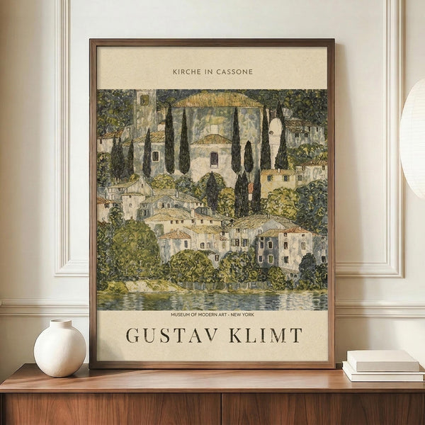 May include: Framed reproduction of Gustav Klimt's "Kirche in Cassone" displayed on a wooden credenza. The artwork shows a Mediterranean hillside village with white buildings, tall cypress trees, and a domed church, rendered in Klimt's distinctive style with rich greens and earth tones. The poster features museum exhibition text above and below the image. A cream ceramic vase and white books sit alongside the brown-framed print against beige walls with white molding.