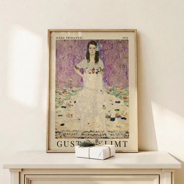 May include: Framed art print showing a woman in a flowing white dress with floral embroidery against a purple patterned background. The artwork features Klimt's distinctive decorative style with colorful geometric patterns on the floor. Displayed in a light wood frame on a cream-colored wooden shelf next to a small white wrapped gift with evergreen sprig.