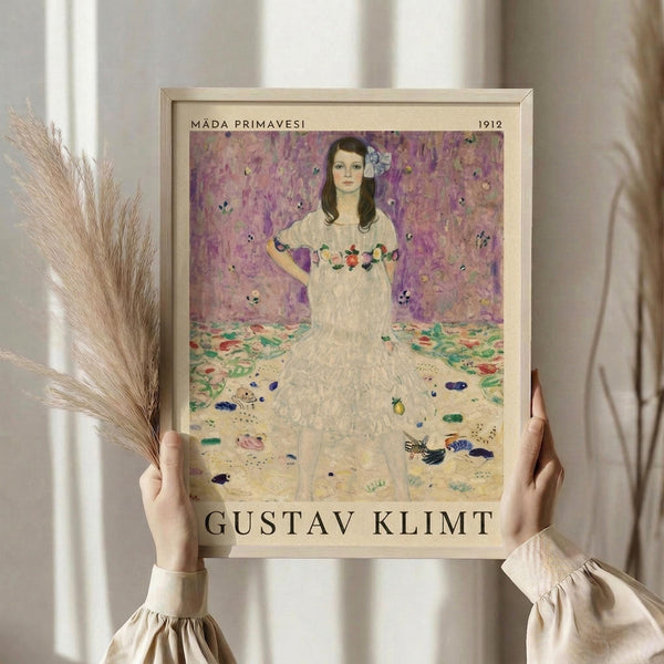 May include: Hands holding framed art print of young girl in white dress with floral details against purple background, displayed in light wood frame. Person wears beige ruffled sleeves, holds dried wheat stems, standing by white curtained window in soft natural lighting.
