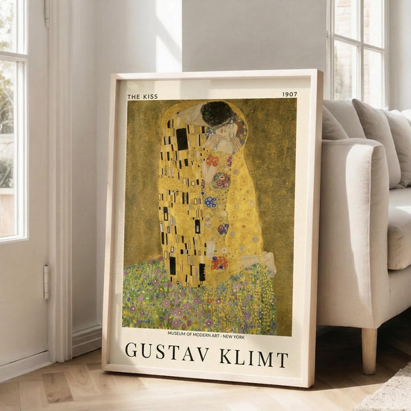 May include: A framed reproduction of Klimt's famous golden artwork showing two embracing figures in ornate robes with geometric patterns and floral motifs, displayed in a light wood frame against a beige armchair in a bright room with white walls and windows.